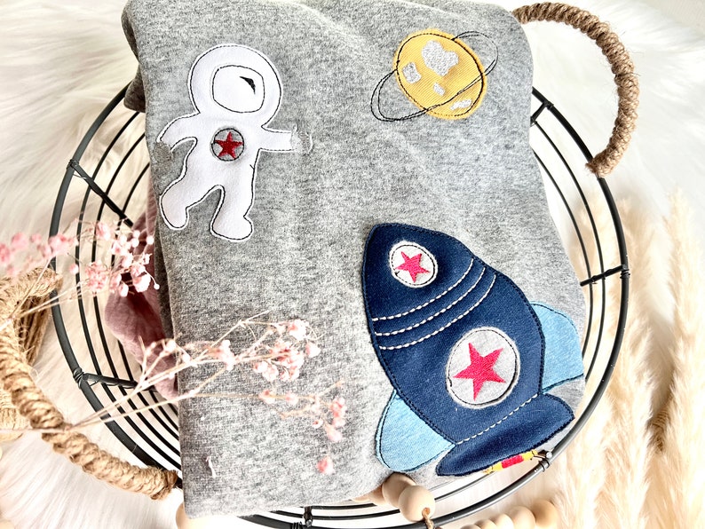 May include: A gray sweatshirt with an astronaut, rocket, and planet applique. The astronaut is white with red and black details. The rocket is blue with red stars and white accents. The planet is yellow with a white heart and a black ring.