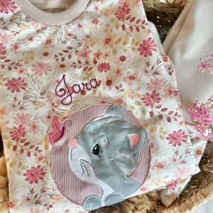 Könnte beinhalten: Ein weißer und rosa floral bedruckter Baby-Strampler mit einer grauen gestickten Katze und dem Namen "Jara" auf der Vorderseite gestickt. Der Strampler befindet sich in einem Weidenkorb mit rosa Blumen.