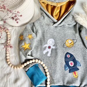 May include: Gray hooded sweatshirt with blue and yellow accents. The sweatshirt features embroidered astronaut, rocket, planet, and star designs.