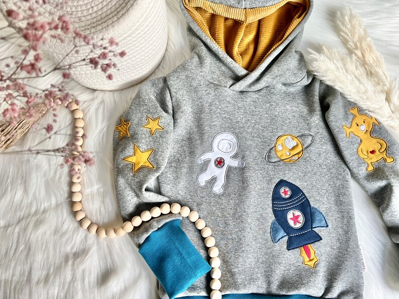 May include: Gray hooded sweatshirt with blue and yellow accents. The sweatshirt features embroidered astronaut, rocket, planet, and star designs.
