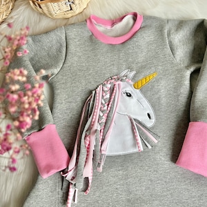 Puede incluir: Vestido de sudadera gris con ribetes rosas y una aplicaci&oacute;n de unicornio blanco con un cuerno amarillo y una crin trenzada rosa y blanca.