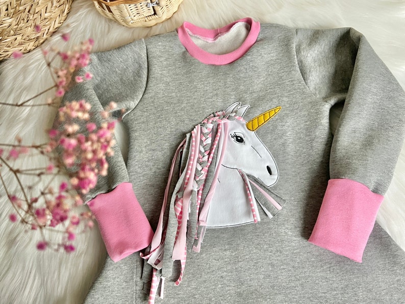 Puede incluir: Vestido de sudadera gris con ribetes rosas y una aplicaci&oacute;n de unicornio blanco con un cuerno amarillo y una crin trenzada rosa y blanca.