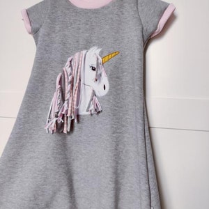 Puede incluir: Un vestido gris con ribetes rosas y una aplicaci&oacute;n de unicornio blanco con un cuerno amarillo y flecos rosas y blancos.
