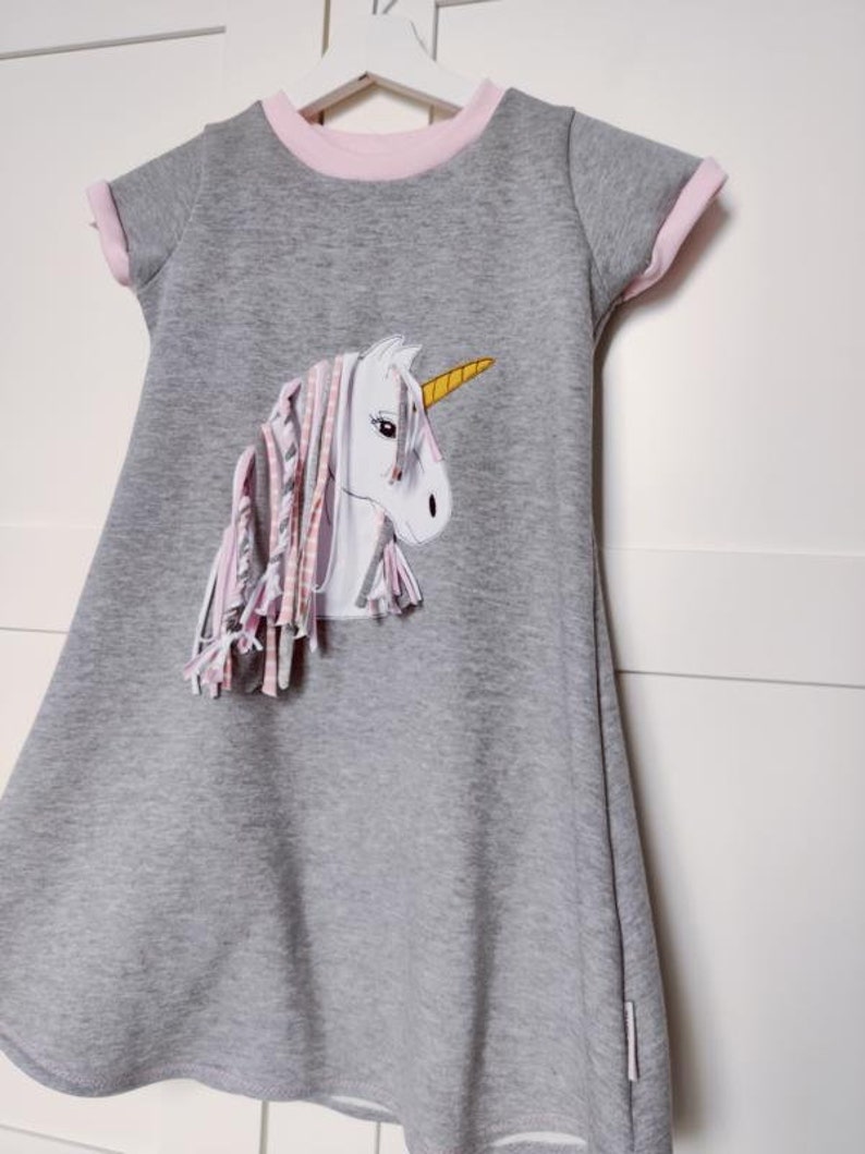 Puede incluir: Un vestido gris con ribetes rosas y una aplicaci&oacute;n de unicornio blanco con un cuerno amarillo y flecos rosas y blancos.
