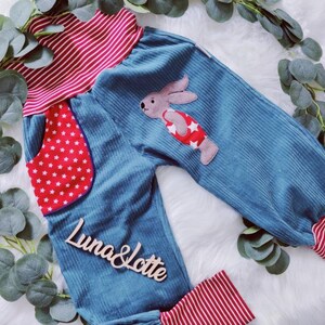 May include: Blue corduroy pants with red and white striped waistband and cuffs. The pants have a red pocket with white stars and a bunny applique on the front. The pants have the text "Luna & Lotte" on the front.