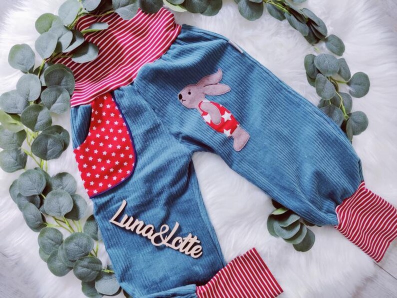May include: Blue corduroy pants with red and white striped waistband and cuffs. The pants have a red pocket with white stars and a bunny applique on the front. The pants have the text "Luna & Lotte" on the front.