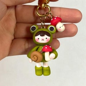 Op de afbeelding: Een grillige sleutelhanger met een miniatuur kikkerpersonage in een groene outfit, met een paddenstoel en een slak. De sleutelhanger heeft een goudkleurige sluiting en een extra paddenstoelbedel. De kikker heeft een verdrietige uitdrukking.