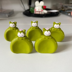 Könnte beinhalten: Fünf grüne dekorative Figuren mit cartoonartigem Design. Jede Figur hat einen abgerundeten Körper, ein weißes Gesicht und eine kleine rosa Blume auf dem Kopf. Die Figuren sind auf einer weißen Oberfläche angeordnet.