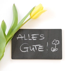 Könnte beinhalten: Eine gelbe Tulpe mit grünen Blättern neben einer kleinen schwarzen Tafel mit der Aufschrift "ALLES GUTE!" in weißer Kreide.
