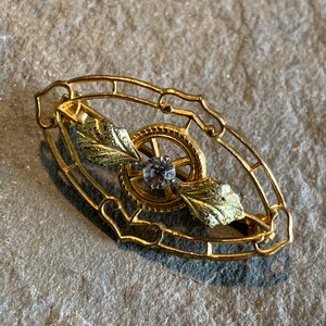May include: A gold-toned brooch with an oval filigree frame. Inside, a gear-like design surrounds a central clear gemstone. Two gold and green leaf accents are on either side of the gear. The brooch is likely vintage or antique.