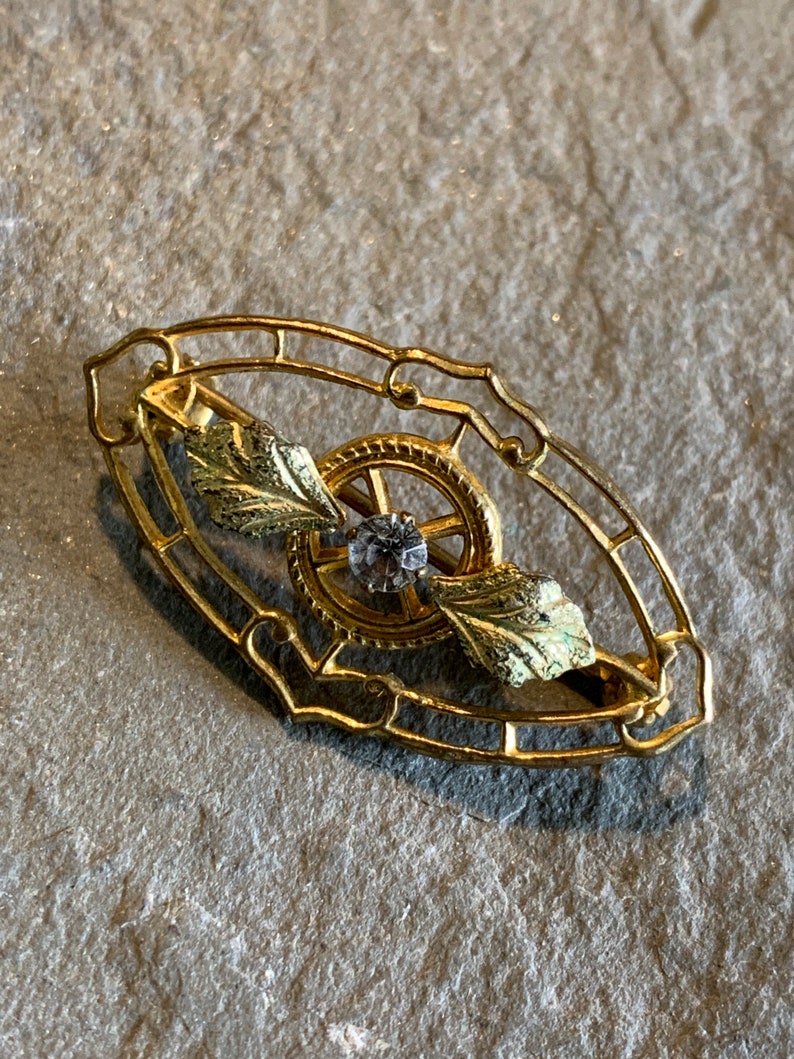 May include: A gold-toned brooch with an oval filigree frame. Inside, a gear-like design surrounds a central clear gemstone. Two gold and green leaf accents are on either side of the gear. The brooch is likely vintage or antique.