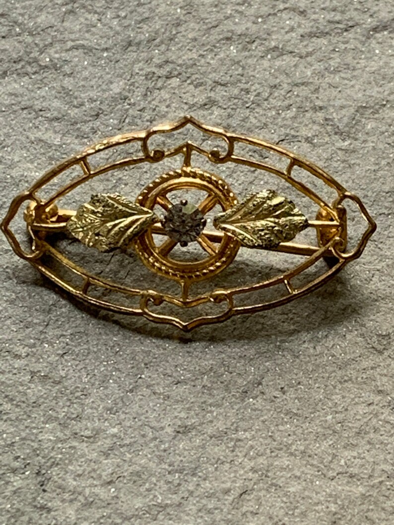 May include: A gold-toned brooch with an oval filigree design. The brooch features a central circular element with a small, dark gemstone and two leaf-shaped accents. The filigree frame has decorative curves.
