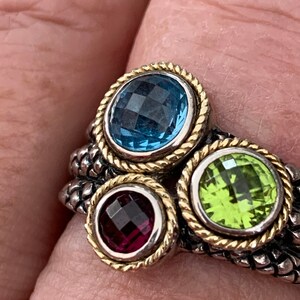 May include: A silver ring with three gemstones. The ring has a textured band and each gemstone is set in a gold bezel. The gemstones are blue, green, and purple.