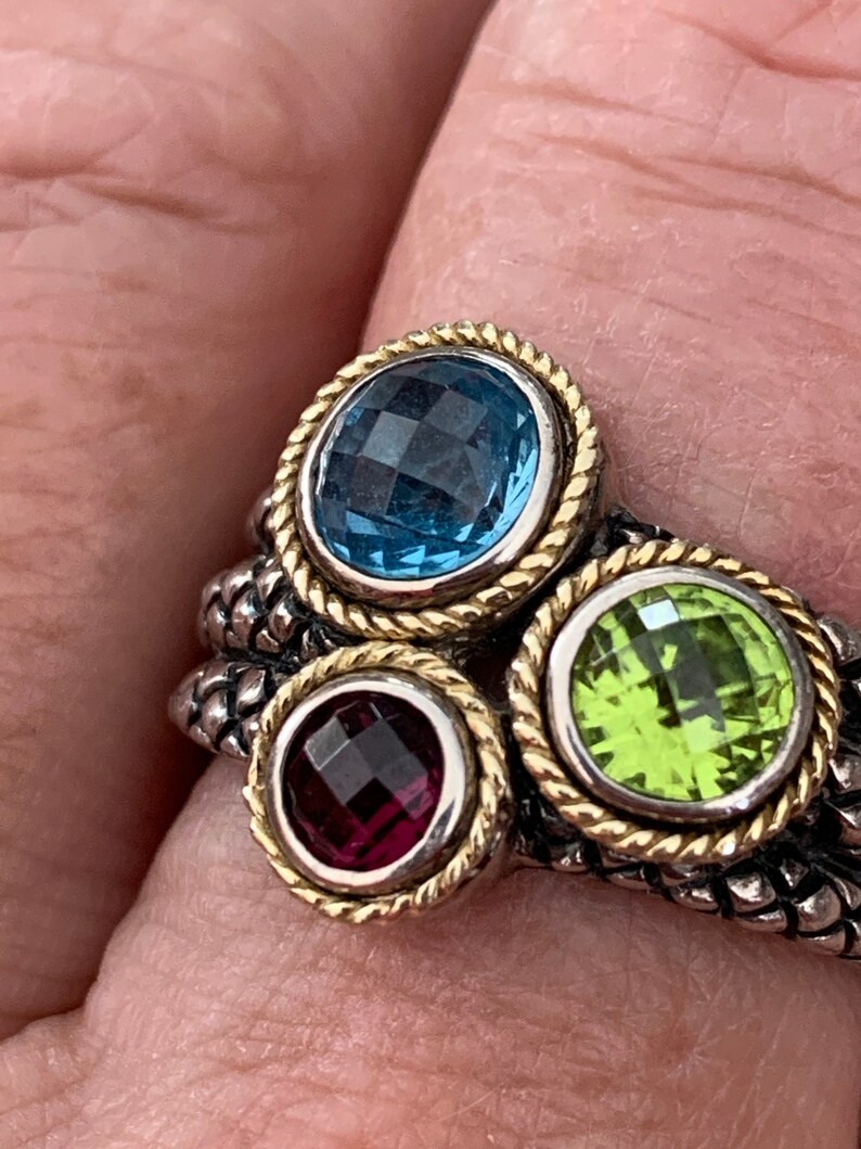 May include: A silver ring with three gemstones. The ring has a textured band and each gemstone is set in a gold bezel. The gemstones are blue, green, and purple.