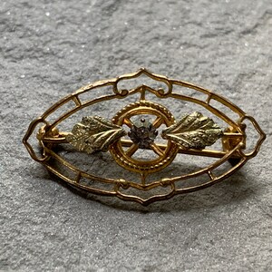 May include: A gold-toned brooch with an oval filigree design. The brooch features a central circular element with a small, round gemstone and two leaf-shaped accents. The pin is secured with a clasp on the back.