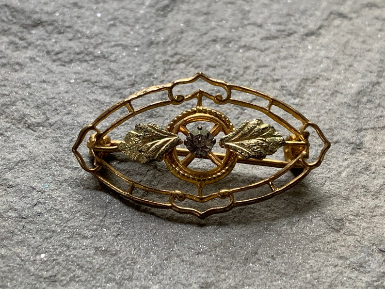 May include: A gold-toned brooch with an oval filigree design. The brooch features a central circular element with a small, round gemstone and two leaf-shaped accents. The pin is secured with a clasp on the back.