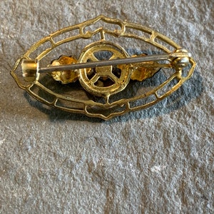 May include: A vintage gold-toned brooch with an oval filigree design. The brooch features a central circular element and two small floral accents. The pin is secured with a clasp on the back. The brooch is likely made of metal.