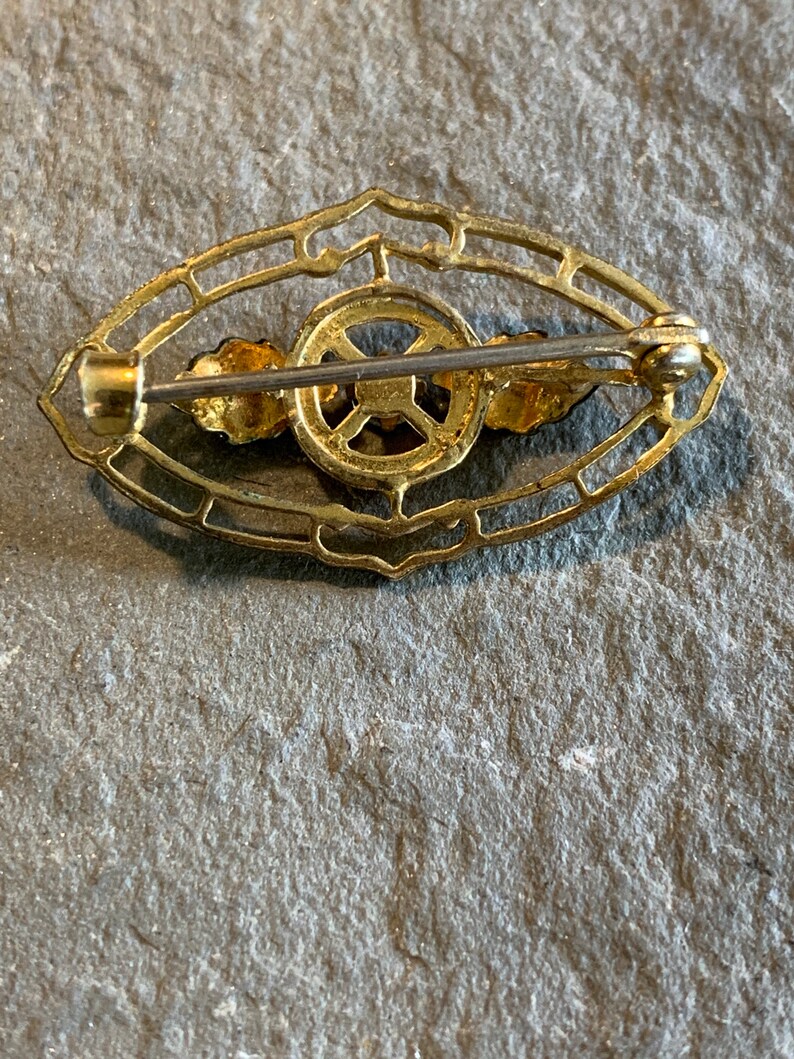 May include: A vintage gold-toned brooch with an oval filigree design. The brooch features a central circular element and two small floral accents. The pin is secured with a clasp on the back. The brooch is likely made of metal.