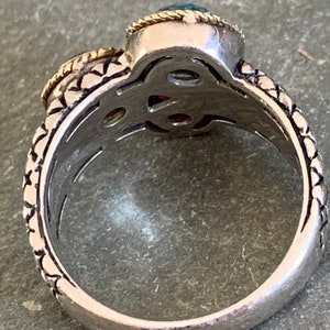 May include: A silver ring with a detailed design and two gemstones. The larger gemstone is blue and the smaller gemstone is red. The ring is set with gold accents.