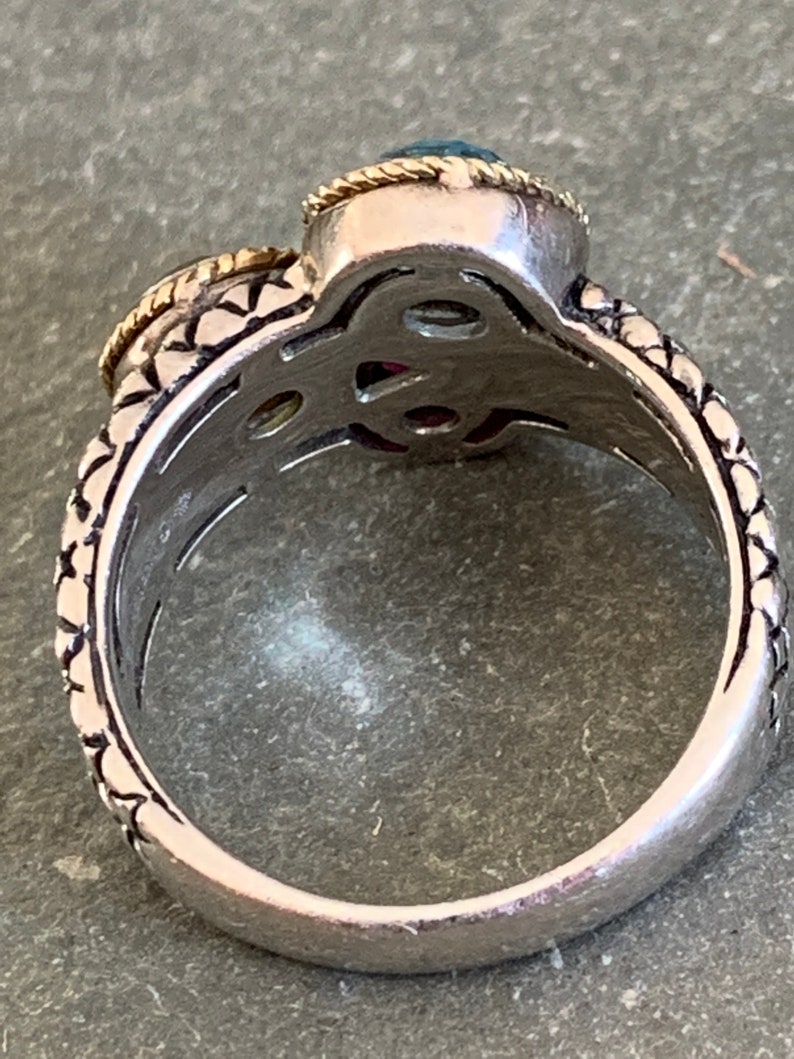 May include: A silver ring with a detailed design and two gemstones. The larger gemstone is blue and the smaller gemstone is red. The ring is set with gold accents.