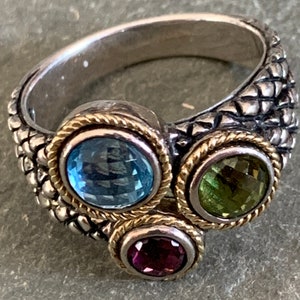 May include: A silver ring with a textured band and three gemstones. The gemstones are a light blue topaz, a green peridot, and a purple amethyst.