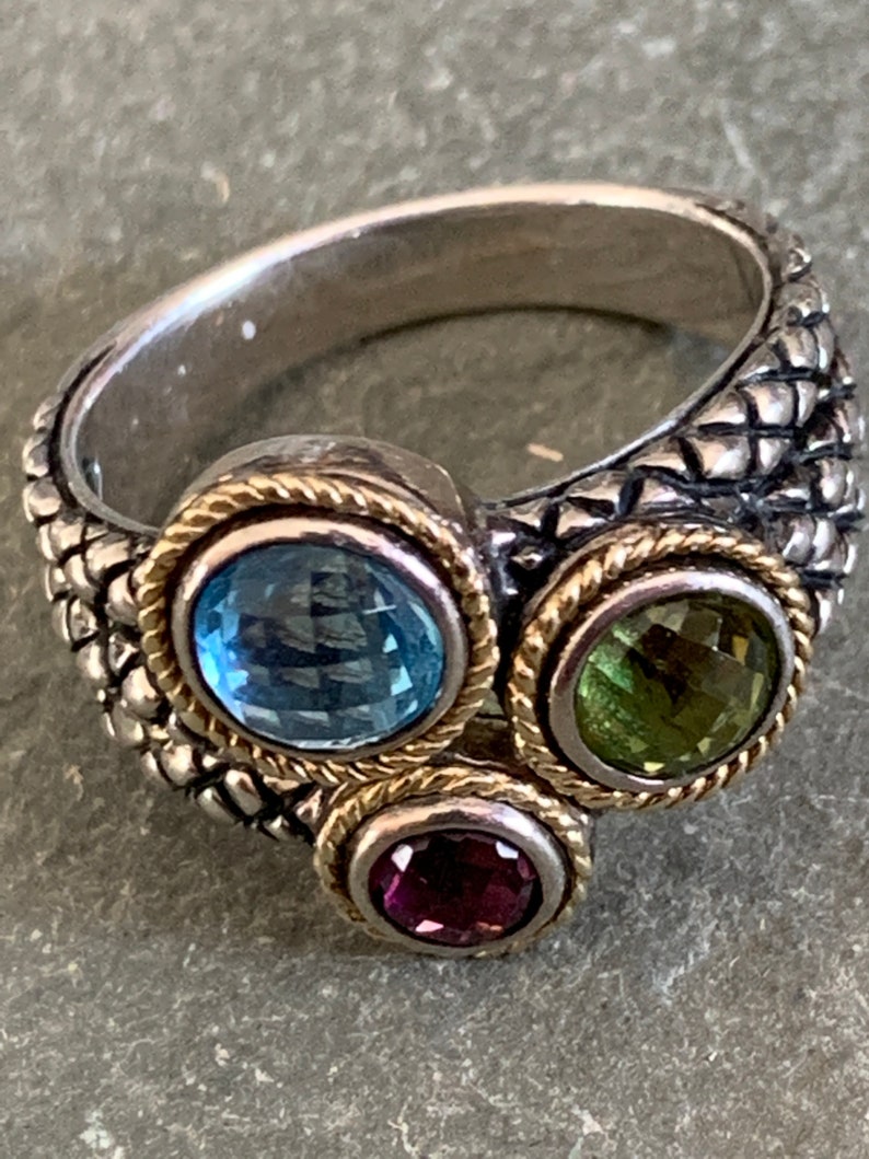 May include: A silver ring with a textured band and three gemstones. The gemstones are a light blue topaz, a green peridot, and a purple amethyst.