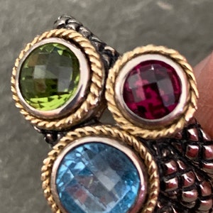 May include: A silver and gold ring with three gemstones. The ring has a rope-like design around the stones. The stones are green, red, and blue.
