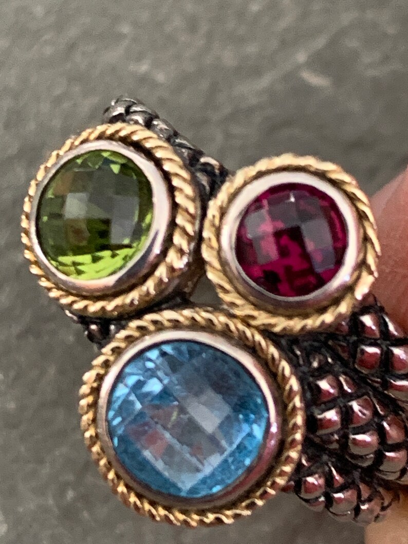 May include: A silver and gold ring with three gemstones. The ring has a rope-like design around the stones. The stones are green, red, and blue.