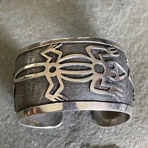 May include: A wide silver cuff bracelet with a textured background and a stylized frog design. The frog is cut out of the silver, creating a contrasting effect. The bracelet has a polished silver edge.