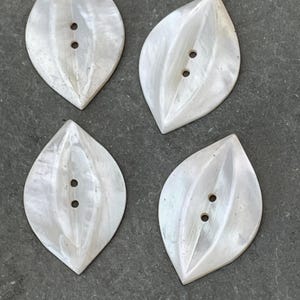 May include: Four pearlescent leaf-shaped buttons with two holes each. The buttons have a subtle, iridescent sheen and a natural, organic form. They are arranged on a dark gray surface.