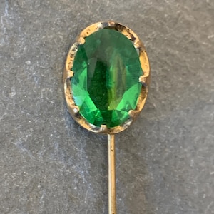 May include: A gold-toned pin with a green glass oval-shaped stone.