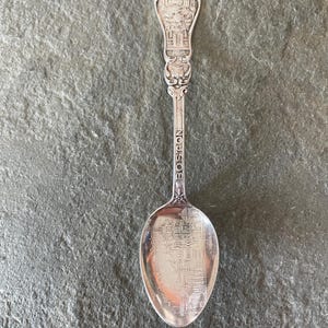 May include: A silver souvenir spoon with the word "BOSTON" engraved on the handle. The spoon's bowl features an etched image of a building and the handle has a detailed architectural design.