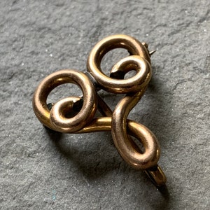 May include: A gold-toned metal brooch in the shape of a stylized heart with a swirl design.