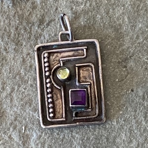 May include: A rectangular silver pendant with an abstract design. The pendant features a small, round, light green stone and a square, dark purple stone. A series of small, round beads are on the left side of the pendant.