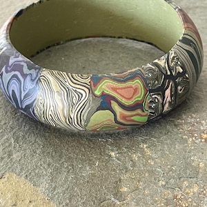 May include: A wide, patterned bangle bracelet. The bracelet features a mix of colors including green, brown, black, and purple. The interior of the bracelet is a solid light green color. The bracelet is resting on a gray stone surface.