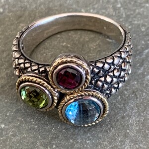 May include: A silver ring with a textured band and three gemstones. The gemstones are a light green, a dark purple, and a light blue.