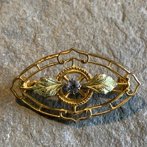 May include: A gold-toned brooch with an oval filigree design. The brooch features a central circular element with a small, round, gray gemstone. Two green and gold leaf accents flank the center. The brooch is set against a gray stone surface.