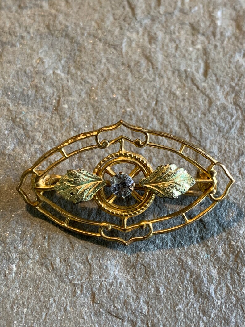 May include: A gold-toned brooch with an oval filigree design. The brooch features a central circular element with a small, round, gray gemstone. Two green and gold leaf accents flank the center. The brooch is set against a gray stone surface.