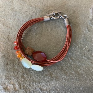 May include: A multi-strand leather bracelet with a silver clasp and colorful gemstone beads. The bracelet features red, orange, yellow, and white beads.