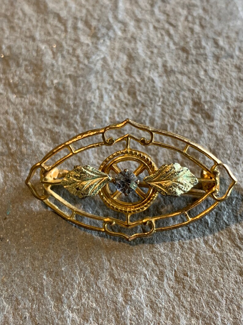 May include: A gold-toned brooch with an oval filigree design. The brooch features a central circular element with a small, dark gemstone and stylized leaf accents. The overall design is ornate and vintage-inspired.