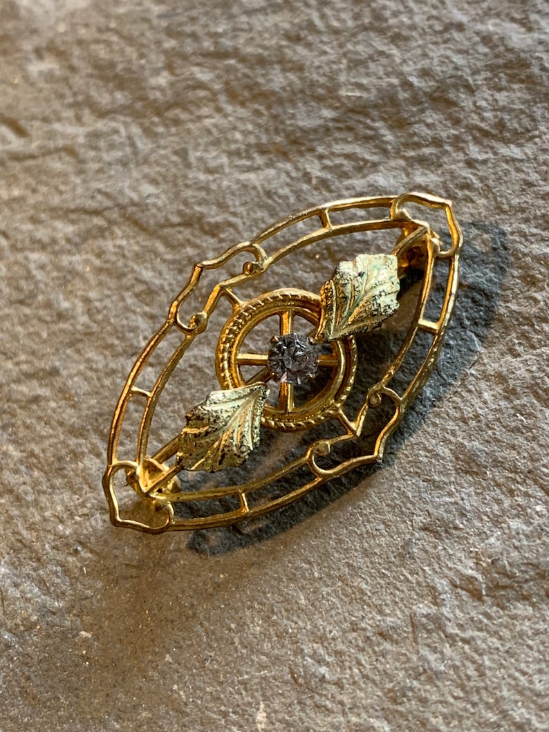 May include: A gold-toned brooch with an oval filigree frame. Inside, a central design features a dark-colored gem surrounded by a wheel-like pattern and two gold-colored leaf accents. The brooch is a vintage style.