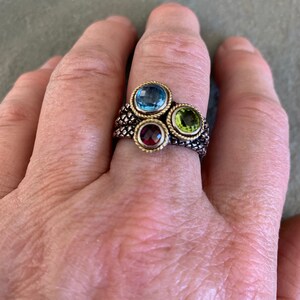 May include: A gold ring with a braided band and four gemstones. The gemstones are blue, green, red, and clear.