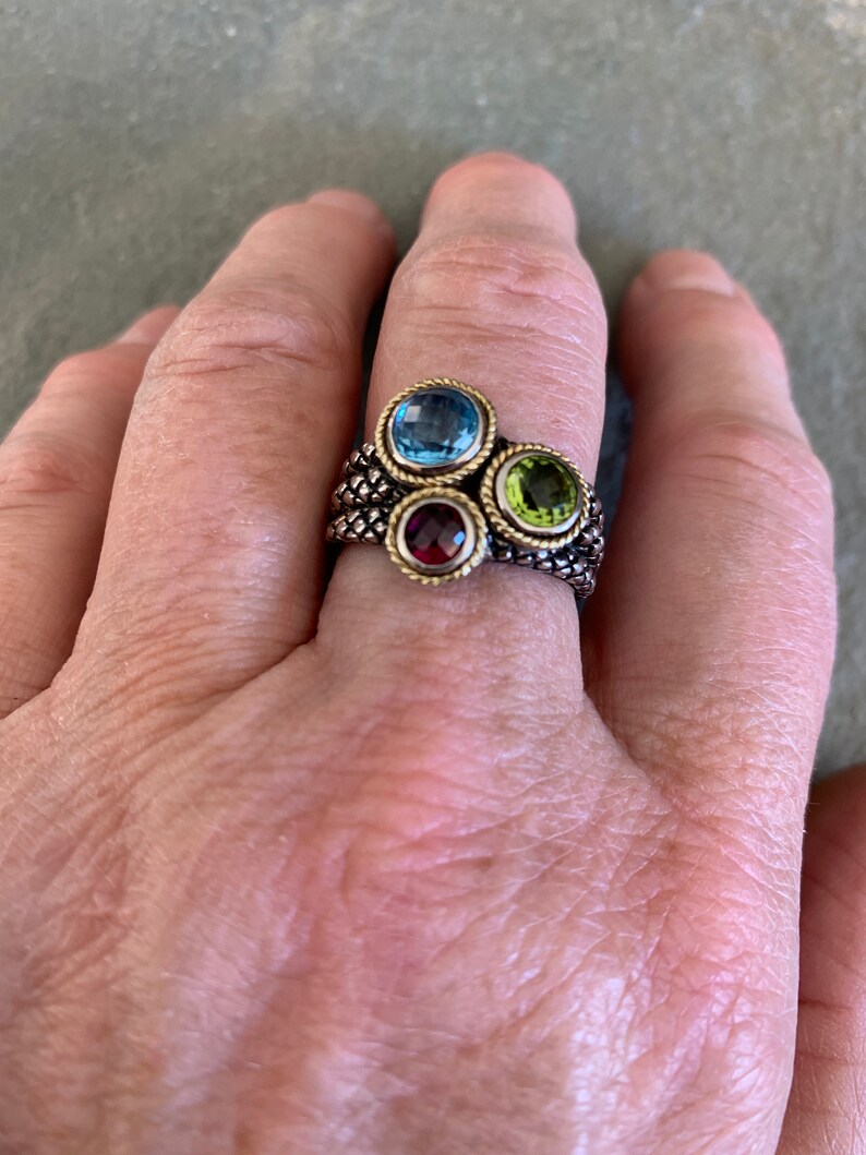 May include: A gold ring with a braided band and four gemstones. The gemstones are blue, green, red, and clear.