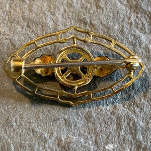May include: A vintage gold-toned brooch with an oval filigree design. The brooch features a central circular element with a cross-like pattern, flanked by two small, stylized wing-like shapes. The pin closure is visible on the back.