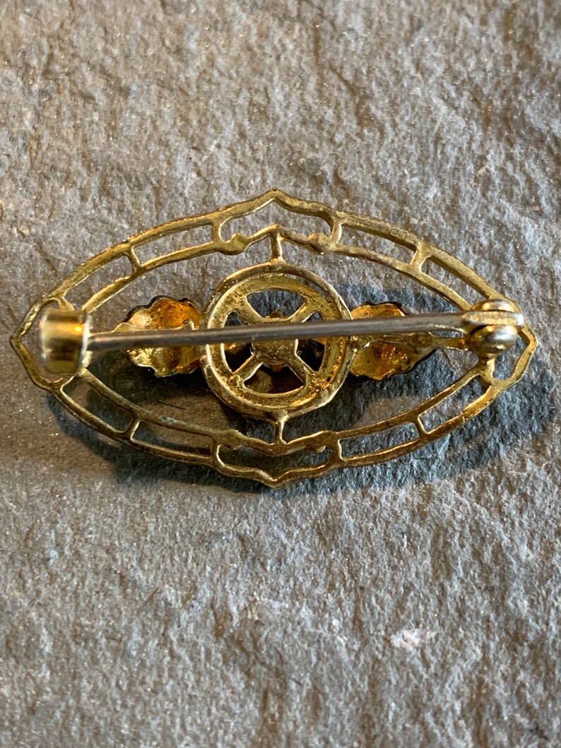 May include: A vintage gold-toned brooch with an oval filigree design. The brooch features a central circular element with a cross-like pattern, flanked by two small, stylized wing-like shapes. The pin closure is visible on the back.