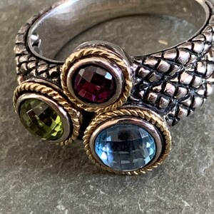 May include: A silver ring with a textured band and three gemstones. The gemstones are a purple amethyst, a green peridot, and a blue topaz. The ring is accented with gold trim.