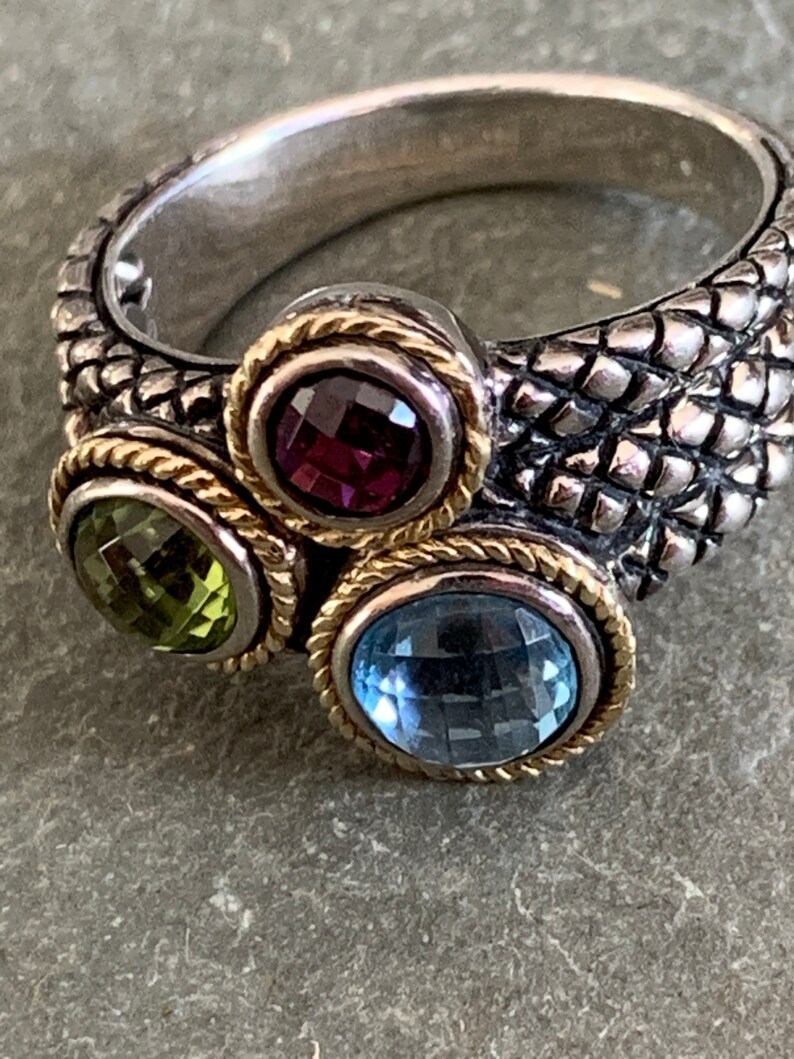 May include: A silver ring with a textured band and three gemstones. The gemstones are a purple amethyst, a green peridot, and a blue topaz. The ring is accented with gold trim.