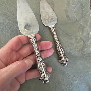 May include: Two silver cake servers with ornate handles. The blades are triangular and show signs of wear. The handles feature a detailed floral design. The servers are displayed on a textured, gray surface.
