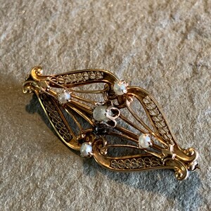 May include: A gold filigree brooch with a delicate design and small white pearls. The brooch is in the shape of a diamond and has a vintage aesthetic.