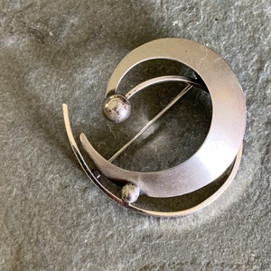 May include: A silver abstract brooch with two round accents. The brooch is shaped like a crescent moon with a smaller curved line intersecting it.